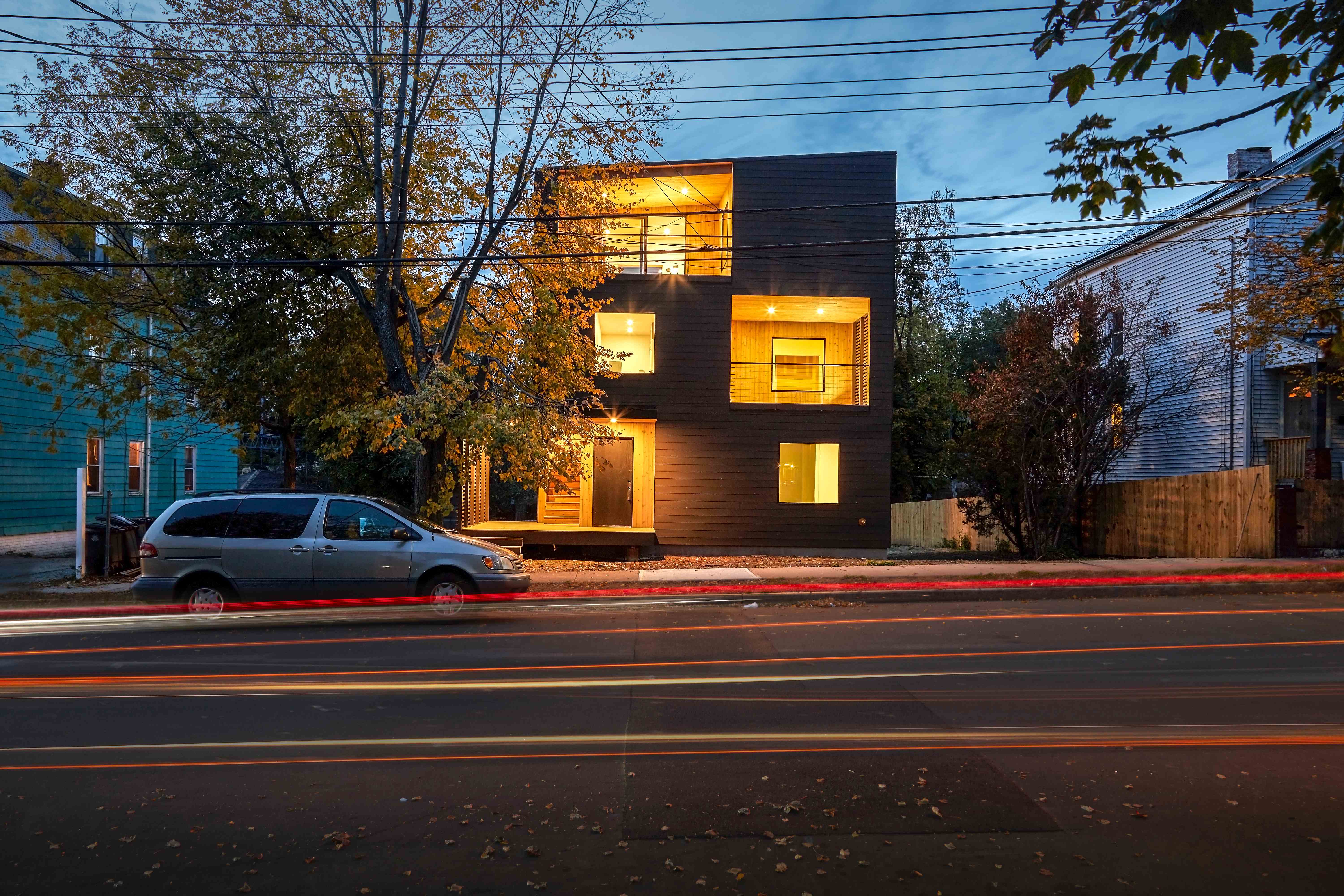 Modern multi-story home with dark cladding and warm interior lighting at dusk with car light trails